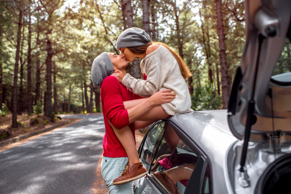 Young and lovely couple in sweates and hats having fun hugging together near the car on the roadside in the pine forest - How to Love a Leo Man